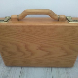 May include: A wooden briefcase with a natural wood finish and brass hardware. The briefcase has a handle on the top and a latch on each side.
