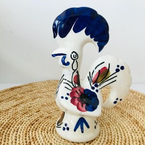 Peut inclure: Figurine de coq en céramique blanche, ornée de motifs floraux bleus, rouges et jaunes. Le coq a une grande crête bleu foncé et des détails peints. Posé sur un tapis tissé. Mesures en cm.