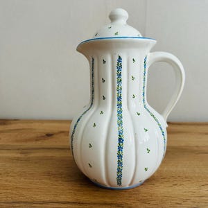 Puede incluir: Jarra de cerámica blanca con tapa, con diseños florales y de hojas azules. La jarra tiene un asa curva y un pico. El diseño incluye rayas azules verticales con pequeños acentos florales azules y verdes. La jarra está sobre una superficie de madera.