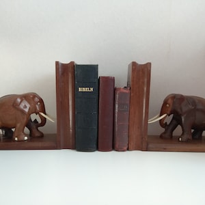 Puede incluir: Un par de sujetalibros de elefante de madera con colmillos de marfil tallados. Los sujetalibros sostienen cuatro libros, uno de los cuales se titula "BIBELN".