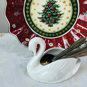 Puede incluir: Porta cubiertos de cerámica blanca con forma de cisne con cucharas de plata en su interior. El cisne está sentado sobre un mantel blanco con un plato de Navidad rojo y verde en el fondo.