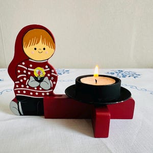 May include: A red wooden candle holder featuring a stylized figure in a hooded outfit with a white dotted pattern. The figure holds a candle, and a lit tea light sits in a black metal holder. The candle holder is on a white tablecloth.