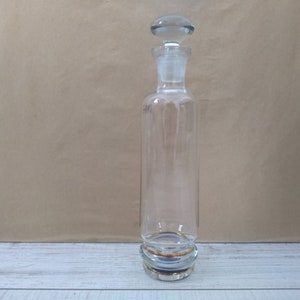 May include: A clear glass bottle with a gold rim and a glass stopper. The bottle is empty and has a textured surface.