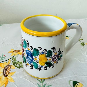 Puede incluir: Taza de cerámica blanca con borde y asa amarillos, adornada con un diseño floral en azul, verde, amarillo y morado. La taza está decorada con un estampado floral pintado a mano. Es un artículo de menaje.