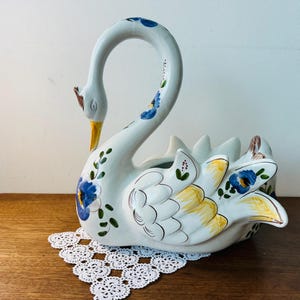 Puede incluir: Una maceta de cisne de cerámica blanca con cuello curvo y parte trasera abierta. El cisne está decorado con motivos florales azules, detalles amarillos y un pico amarillo. Se encuentra sobre un tapete de encaje blanco.