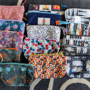 May include: A collection of 13 colourful zippered pouches with various patterns, including geometric designs, dice, bicycles, and paisley. The pouches are made of fabric and have a variety of colours, including blue, green, red, yellow, and white.