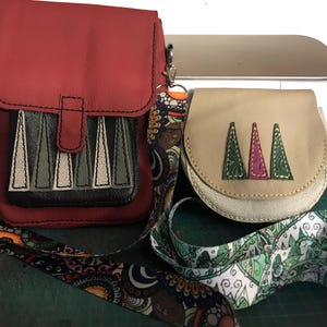 May include: A red rectangular crossbody bag with a black front pocket featuring five triangular appliques, paired with a smaller beige rounded bag featuring three triangular appliques. Both bags have long, patterned fabric shoulder straps.