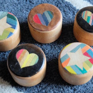 May include: Six small wooden boxes with different colorful designs on the lids. The designs include a star, a heart, and a rainbow.