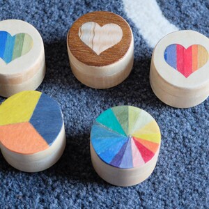 May include: Five wooden boxes with different designs. The boxes are round and have a heart, a rainbow heart, a colour wheel, and two different geometric designs.