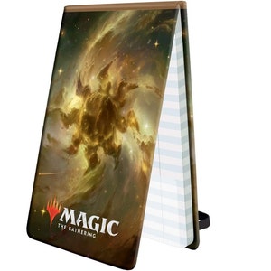 May include: A Magic: The Gathering notebook with a galaxy-themed cover featuring a swirling nebula in shades of gold, brown, and white. The notebook has lined pages and a black elastic closure.