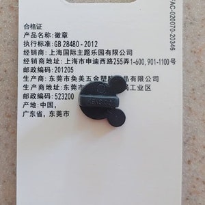 May include: A black and white pin with a Mickey Mouse head design. The pin is attached to a white card with text that includes the words "Disney", "Made in China", "合格证", "产品名称", "执行标准", "经销商", "经销商地址", "邮政编码", "生产商", "生产商地址", "邮政编码", "产地", "广东省", "东莞市", and "岗工业区".