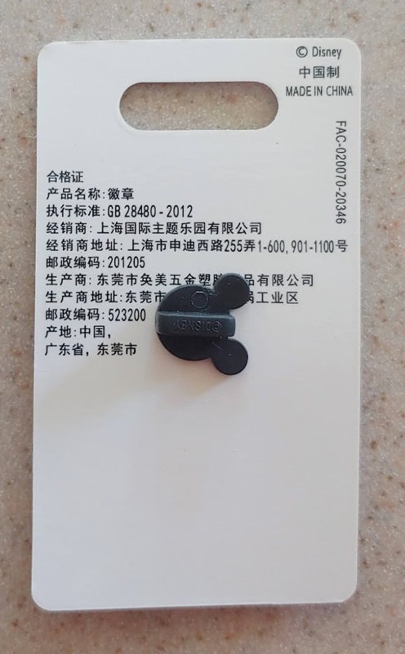 May include: A black and white pin with a Mickey Mouse head design. The pin is attached to a white card with text that includes the words "Disney", "Made in China", "合格证", "产品名称", "执行标准", "经销商", "经销商地址", "邮政编码", "生产商", "生产商地址", "邮政编码", "产地", "广东省", "东莞市", and "岗工业区".