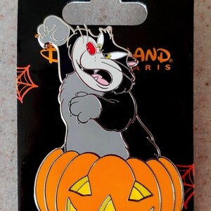 May include: A black and gray cat with a red eye is perched on top of an orange pumpkin with a carved jack-o'-lantern face. The cat is holding its paw up in the air. The background is black with a spider web and stars. The text "Disneyland Paris" is visible in the upper right corner.