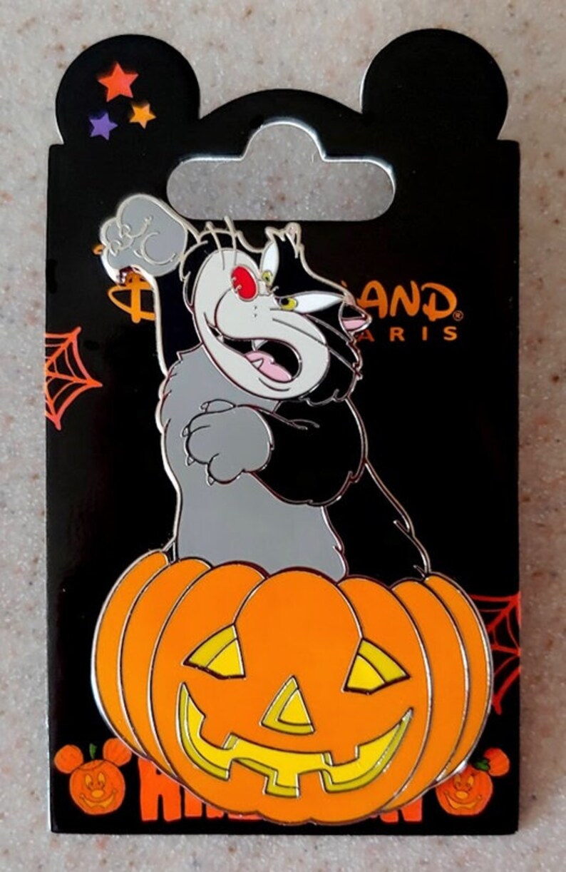 May include: A black and gray cat with a red eye is perched on top of an orange pumpkin with a carved jack-o'-lantern face. The cat is holding its paw up in the air. The background is black with a spider web and stars. The text "Disneyland Paris" is visible in the upper right corner.