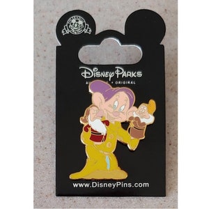 May include: A yellow and purple Disney Parks pin featuring a cartoon depiction of Doc from Snow White and the Seven Dwarfs. Doc is holding a smaller version of himself in his right hand. The pin has the text "Disney Parks" and "www.DisneyPins.com".