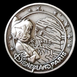 May include: Silver-toned metal coin featuring Peter Pan and a depiction of Skull Rock from the Peter Pan's Flight ride at Disneyland Paris. The coin has a starburst design around the edge and the text "Disneyland Paris".