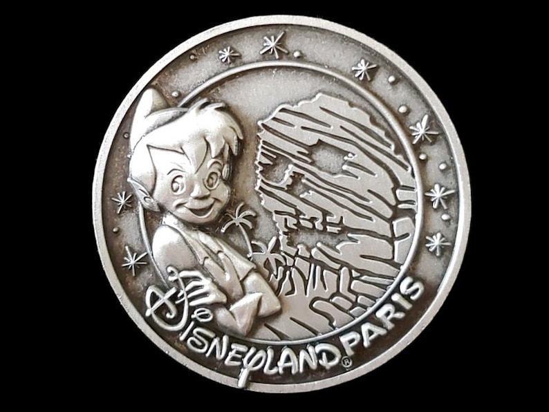May include: Silver-toned metal coin featuring Peter Pan and a depiction of Skull Rock from the Peter Pan's Flight ride at Disneyland Paris. The coin has a starburst design around the edge and the text "Disneyland Paris".