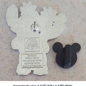 May include: Silver-toned metal pin featuring Stitch from Lilo & Stitch. Stitch is wearing a red Santa hat and holding a gift. The pin is approximately 1 3/4 inches tall and 1 3/8 inches wide. The back of the pin has the Disney logo and "Made in China" printed on it.