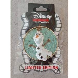 May include: A gold-colored pin with a green background featuring Olaf from the movie Frozen. The background is covered with the word "Olaf" written in a cursive font. The pin is a limited edition Disney Studios Hollywood pin.