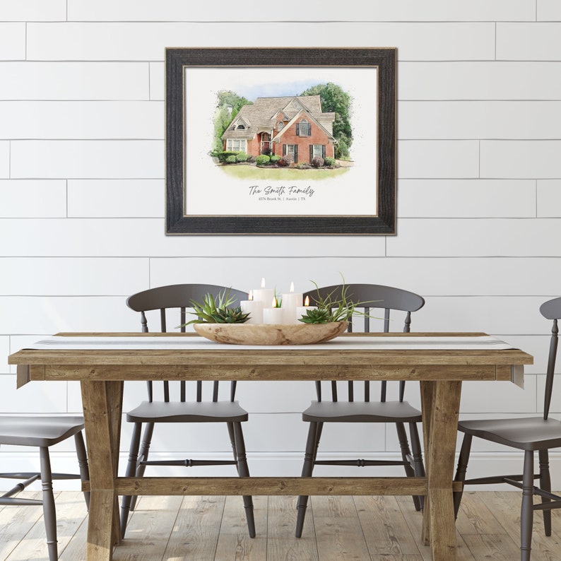 May include: A framed watercolour painting of a red brick house with white trim and a green lawn. The painting is titled "The Smith Family" with an address and date below. The painting is hanging on a white shiplap wall above a wooden table with four chairs.