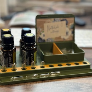 May include: An olive green essential oil organiser with a tray for capsules and a hinged box. Several brown glass bottles with black caps are arranged on the tray. The box has floral accents and the text "Naturally".