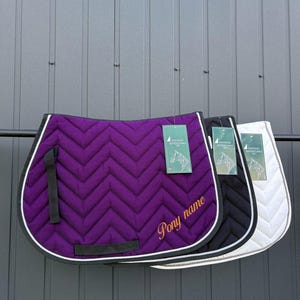 Puede incluir: Tres sudaderos acolchados en morado, negro y blanco, cada uno con patrón de chevrón, ribete negro y borde de cuerda blanca. El sudadero morado muestra el texto bordado en oro Pony name y una etiqueta verde Outdoor Adventures.