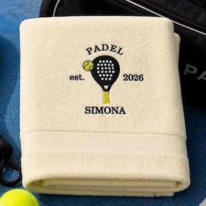 Peut inclure: Une serviette de couleur crème avec une broderie noire indiquant PADEL est. 2026 et SIMONA, accompagnée d'un motif représentant une raquette de padel noire et une balle jaune. La serviette est pliée près d'une raquette et de balles de padel.
