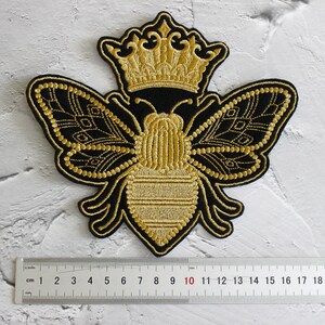 May include: A gold embroidered bee with a crown on its head. The bee is black with gold accents and has wings that look like butterfly wings. The bee is about 10 cm wide.