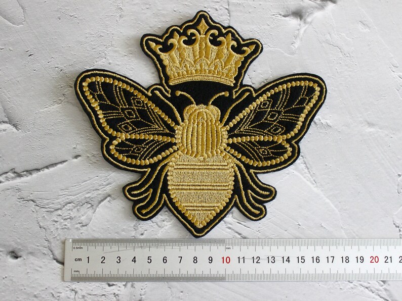 May include: A gold embroidered bee with a crown on its head. The bee is black with gold accents and has wings that look like butterfly wings. The bee is about 10 cm wide.