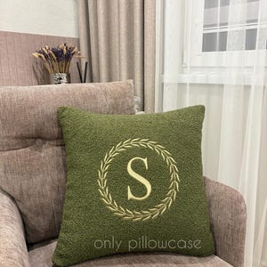 May include: A square olive green textured pillowcase featuring a central embroidered cream-coloured letter S enclosed within a circular laurel wreath design. The text "only pillowcase" is embroidered in small cream letters at the bottom centre.