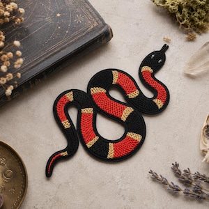 May include: A snake-shaped embroidered patch featuring a repeating pattern of black, bright red, and thin yellow bands. The snake is depicted in a coiled, S-shaped position with a rounded head, visible stitching texture, and a black border outline.