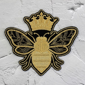 May include: A gold embroidered bee with a crown on its head. The bee is black with gold details and is on a white background.