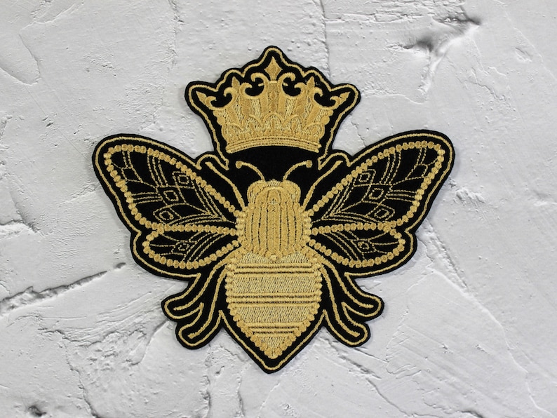 May include: A gold embroidered bee with a crown on its head. The bee is black with gold details and is on a white background.
