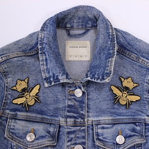 May include: A blue denim jacket with two gold bee patches, each with a crown on top. The jacket has a label that says "DENIM WEAR".