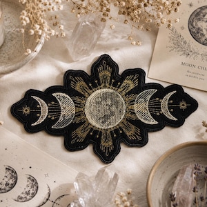 May include: A black embroidered patch featuring a central full moon surrounded by radiating golden sun rays and a series of crescent moon phases on both sides. The design is intricate with metallic gold and white thread detailing on black fabric.