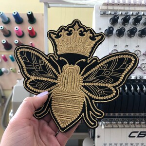 May include: A gold embroidered bee with a crown on its head. The bee is on a black fabric background.