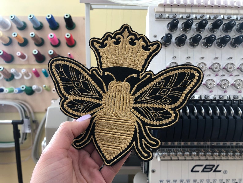 May include: A gold embroidered bee with a crown on its head. The bee is on a black fabric background.