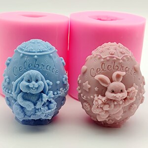 Könnte beinhalten: Zwei Silikonformen zum Herstellen von Osterkerzen in Eiform. Die Formen sind rosa und jede zeigt ein blaues oder rosa Kaninchen, das einen Korb mit dem Wort "Celebrate" auf dem Ei hält.