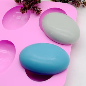 May include: A pink silicone mould with six oval cavities, holding two oval soaps, one light blue and one grey. The mould is designed for soap making or crafting, with a smooth, clean appearance.