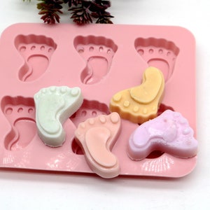 May include: Pink silicone mold with six baby footprint shapes.  Four of the shapes are filled with colorful soap in shades of pink, white, and yellow.