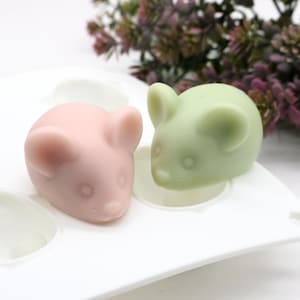 Puede incluir: Un molde de silicona blanco con dos cavidades en forma de ratón. Una cavidad está llena de un jabón en forma de ratón rosa y la otra cavidad está llena de un jabón en forma de ratón verde.