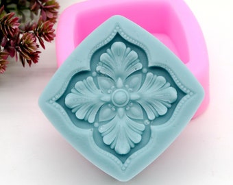 Könnte beinhalten: Eine hellblaue Silikonform mit einem dekorativen Blumenmuster. Die Form ist quadratisch mit abgerundeten Ecken. Die Form liegt auf einer rosa Silikonform.