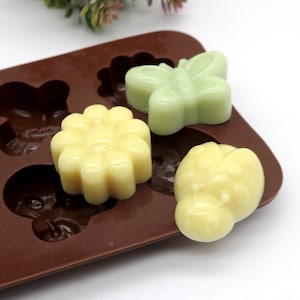 Könnte beinhalten: Eine braune Silikonform mit verschiedenen Tierformen, darunter ein Schmetterling, ein Marienkäfer und eine Blume. Die Form ist mit weißer und grüner Seife gefüllt.