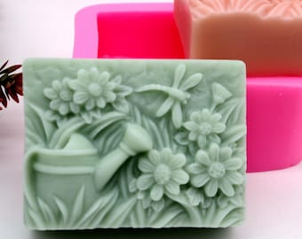 May include: A green soap bar with a detailed design of a watering can, flowers, and a dragonfly. The soap bar is in a pink silicone mold.