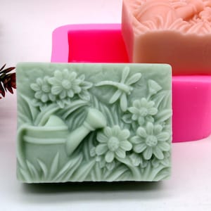 Könnte beinhalten: Eine grüne Seife mit einem detaillierten Design einer Gießkanne, Blumen und einer Libelle. Die Seife befindet sich in einer rosa Silikonform.