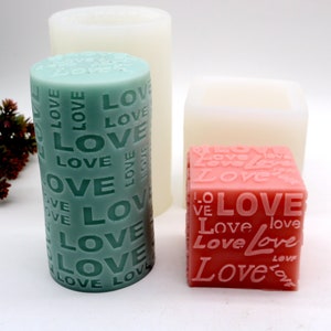 May include: Three silicone molds for making candles. One mold is a square, one is a cylinder, and one is a cylinder with the word "LOVE" repeated all over it. The square mold is white, the cylinder molds are white on the outside and green and pink on the inside.