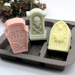 Puede incluir: Un molde de silicona para hacer jabones con forma de lápidas con temática de Halloween. El molde tiene tres cavidades con forma de lápidas. Una lápida tiene una calavera y huesos cruzados, otra tiene la palabra "RIP" y una telaraña, y la tercera tiene una araña. El molde es gris y las barras de jabón son rosas, verdes y amarillas.