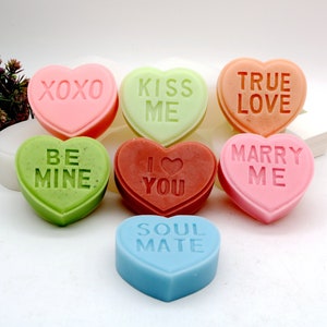 Puede incluir: Ocho jabones en forma de corazón con diferentes mensajes románticos. Los jabones son de varios colores, incluyendo rosa, verde, rojo, naranja y azul. Los mensajes en los jabones incluyen "XOXO", "Kiss Me", "True Love", "Be Mine", "I Love You", "Marry Me", y "Soul Mate".