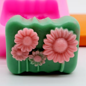 Puede incluir: Una pastilla de jab&oacute;n verde con tres flores de margarita rosas en la parte superior. La pastilla de jab&oacute;n est&aacute; en un molde de silicona.