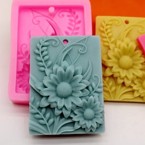 Puede incluir: Un molde de silicona para hacer jabón o fundir cera. El molde tiene forma rectangular con un diseño floral con dos margaritas grandes y hojas. El molde es rosa y el diseño es de color azul claro.
